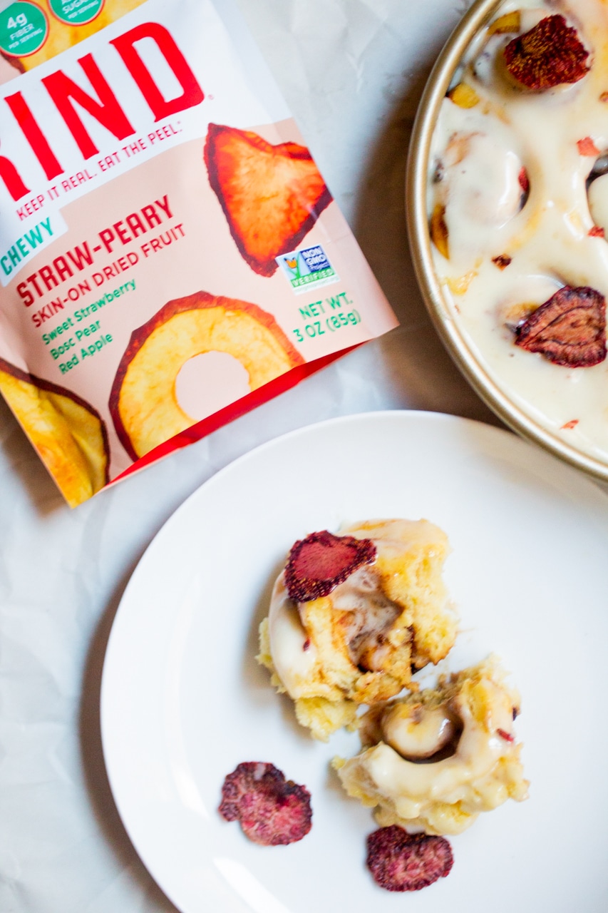 A cinnamon roll with freeze-dried strawberries on a white plate next to a bag of RIND dried strawberries.