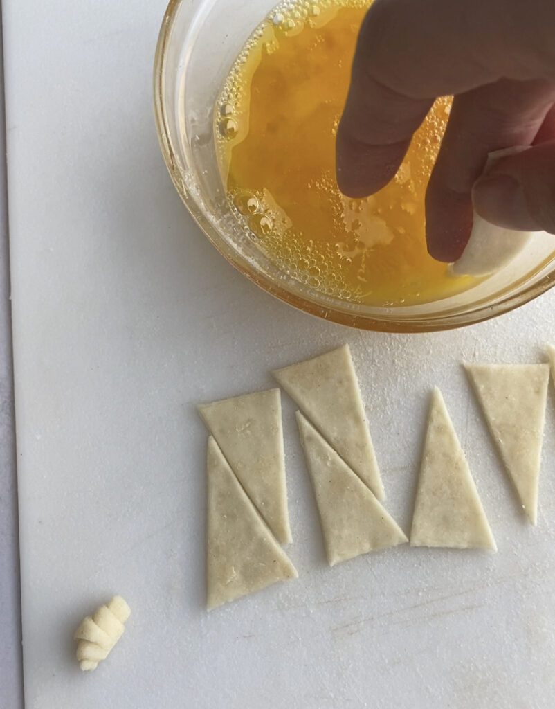 A bunch of triangles of puff pastry next to a bowl of egg wash.