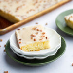 square of white Texas sheet cake on a white plate with a sheet cake in the background.