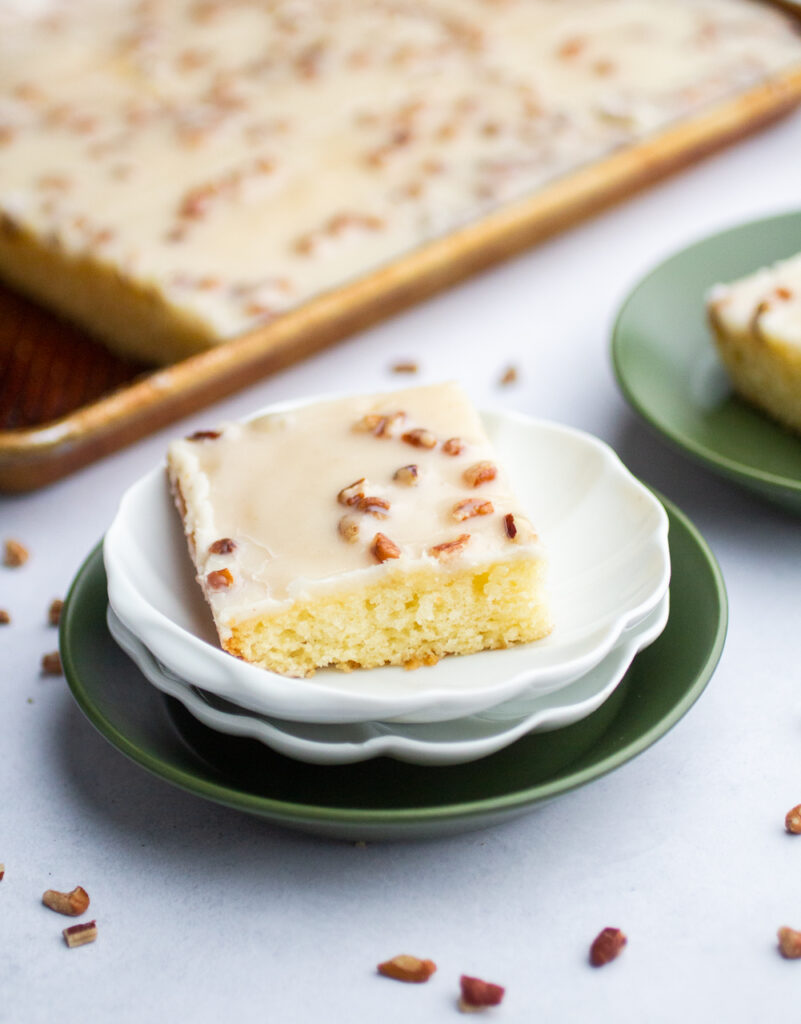 square of white Texas sheet cake on a white plate with a sheet cake in the background.