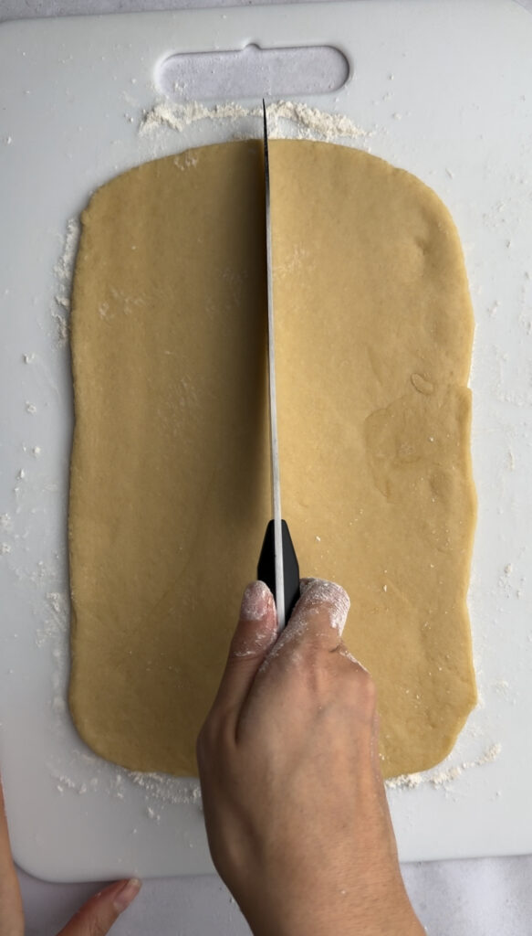 Dough rolled out on a cutting board being cut in half by a knife.