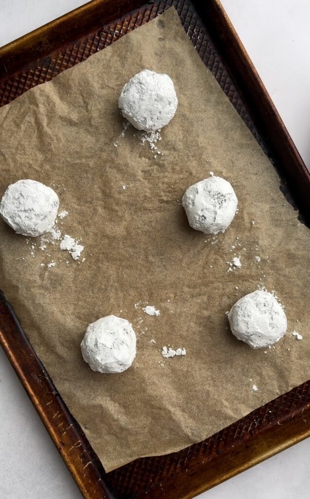Balls of cookie dough covered in powdered sugar on a parchment-lined baking sheet.