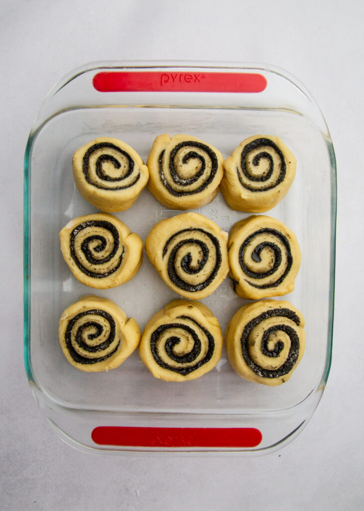 9 black sesame sweet rolls rising in a square glass pan.