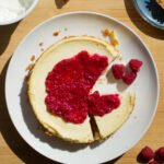 A cheesecake topped with a raspberry compote with a slice missing on a white plate.