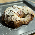 An almond croissant with powdered sugar on a gray baking pan.
