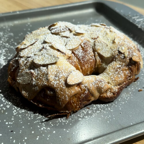 An almond croissant with powdered sugar on a gray baking pan.