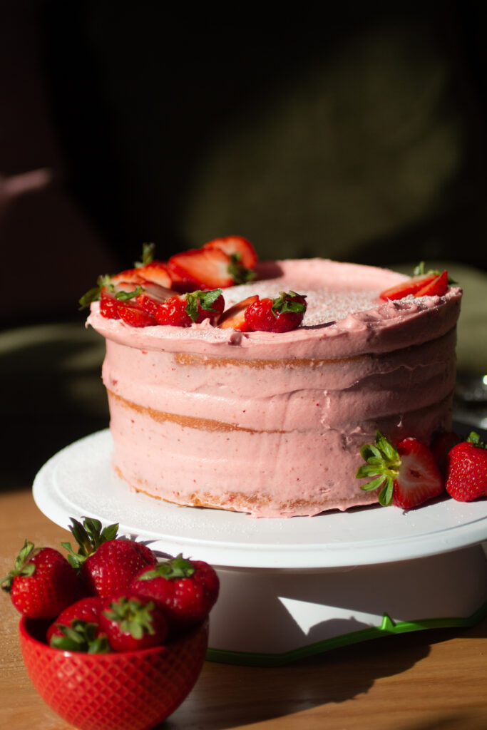 strawberry cake on a white cake stand with a bowl of strawberries.