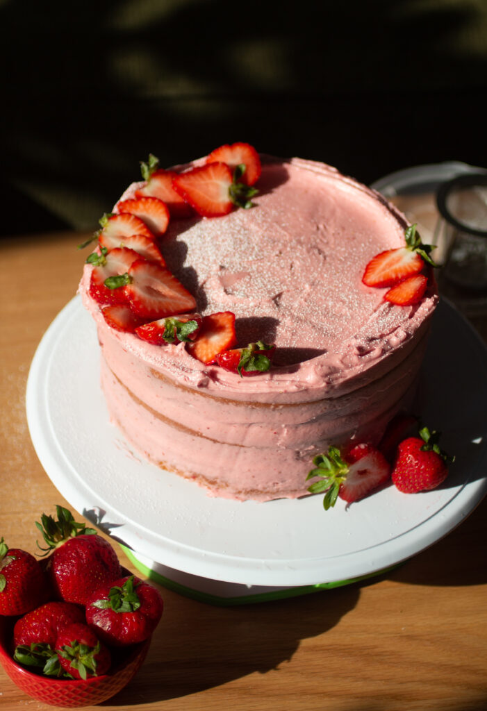 a pink strawberry cake on a white cake stand next to a cup of strawberries.