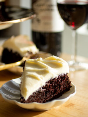 A slice of chocolate cake with cream cheese frosting on a white plate with a glass of red wine in the background.