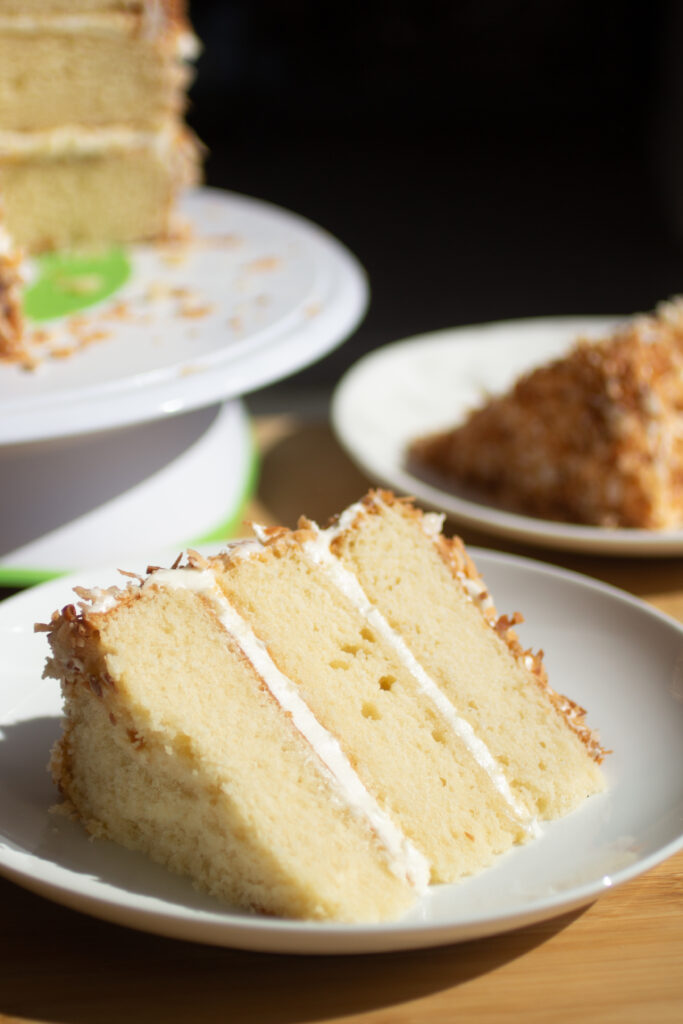 A three layer slice of white cake with vanilla frosting and toasted coconut.