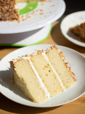 A three layer slice of white cake with vanilla frosting and toasted coconut.