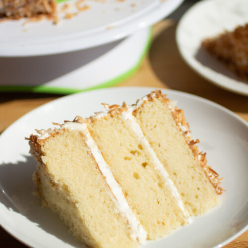 A three layer slice of white cake with vanilla frosting and toasted coconut.