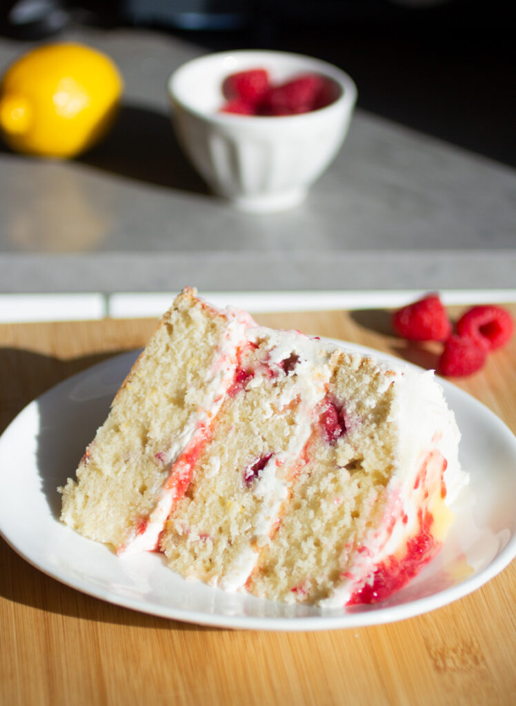 A three layer slice of vanilla cake with raspberries and white frosting on a white plate.