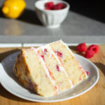 A three layer slice of vanilla cake with raspberries and white frosting on a white plate.