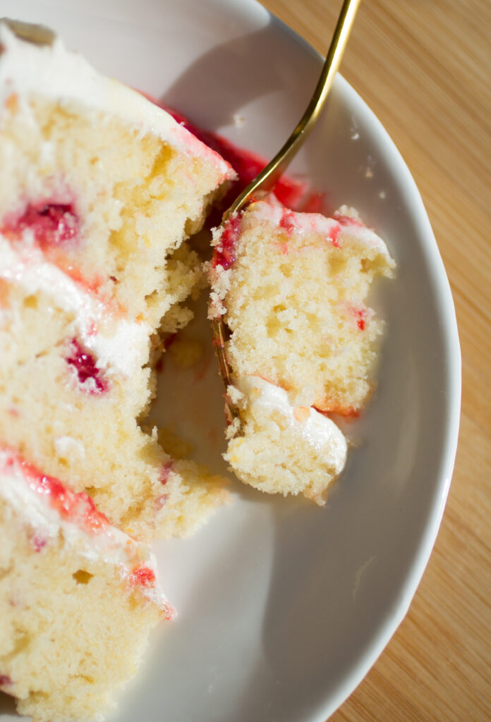 A fork taking a bite out of a three layer slice of vanilla cake with raspberries and white frosting on a white plate.