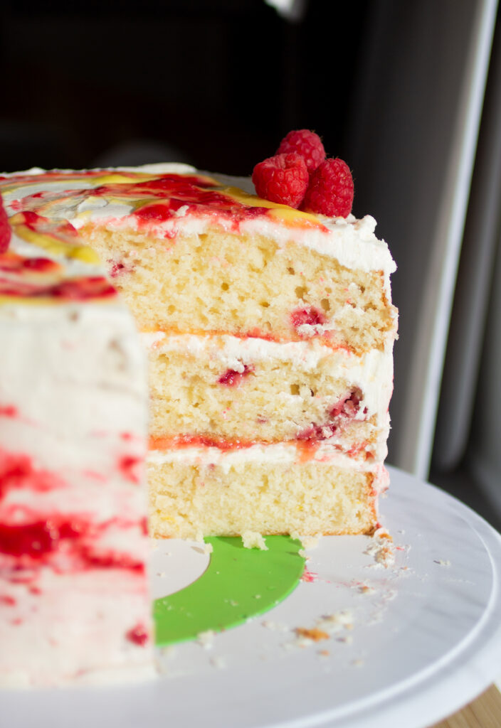 A three layer vanilla cake with raspberries and lemon curd on a cake stand with a slice cut out to show the interior.