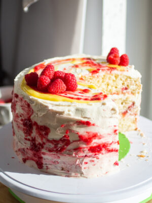 A three layer vanilla cake with raspberries and lemon curd on a cake stand with a slice cut out to show the interior.
