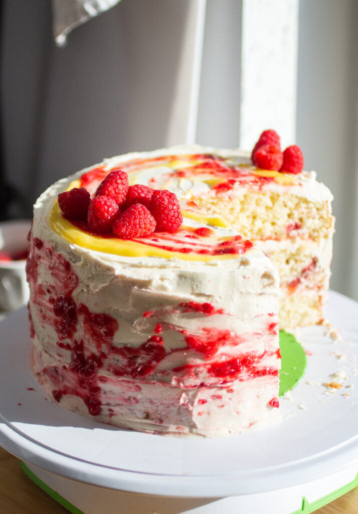 A three layer vanilla cake with raspberries and lemon curd on a cake stand with a slice cut out to show the interior.