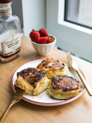 Three thick pancakes on a white plate with a red rim next to a bowl of strawberries and a jar of maple syrup.
