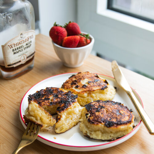 Three thick pancakes on a white plate with a red rim next to a bowl of strawberries and a jar of maple syrup.