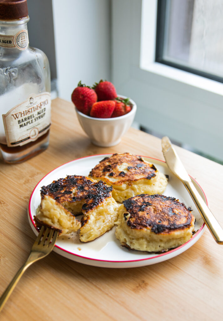 Three thick pancakes on a white plate with a red rim next to a bowl of strawberries and a jar of maple syrup.