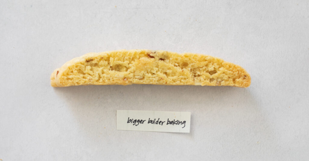 A pice of biscotti on a gray background with the label "Bigger Bolder Baking."