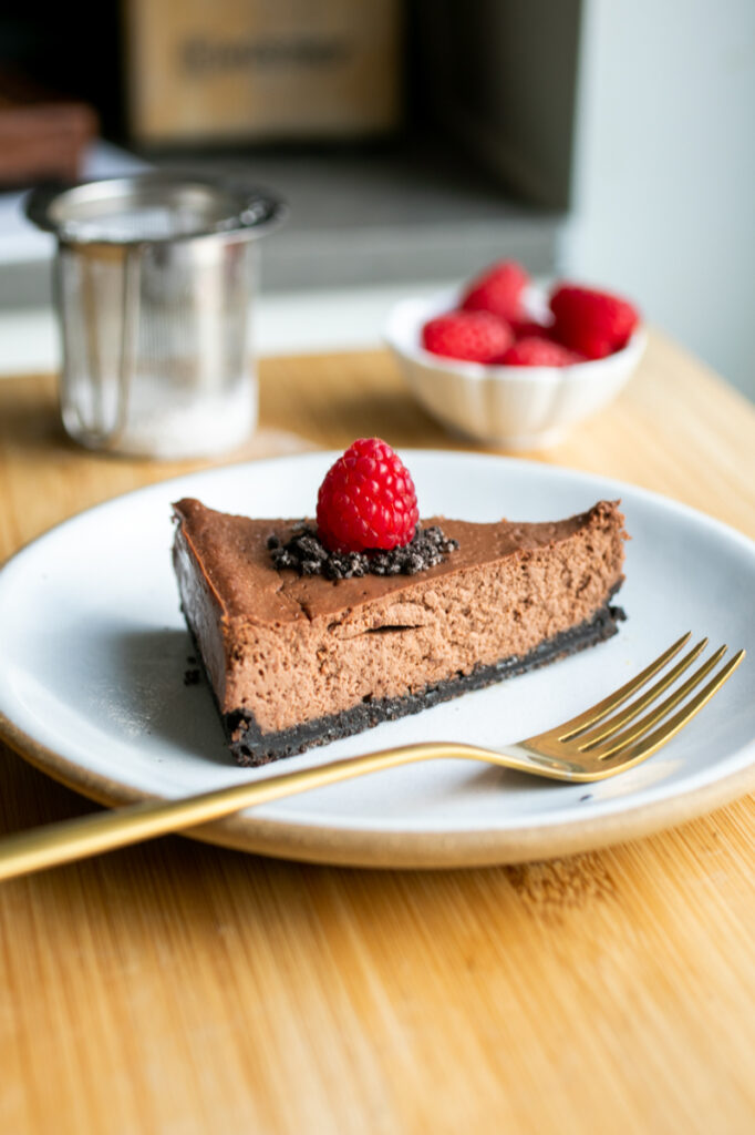 A slice of chocolate cheesecake on a white plate topped with a raspberry.