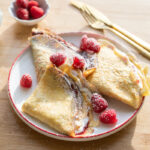 Three crepes with raspberries on a white plate.