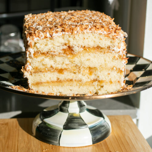 A four-layer coconut cake covered in toasted coconut on a black and white cake stand with a slice taken out to show the cross-section.