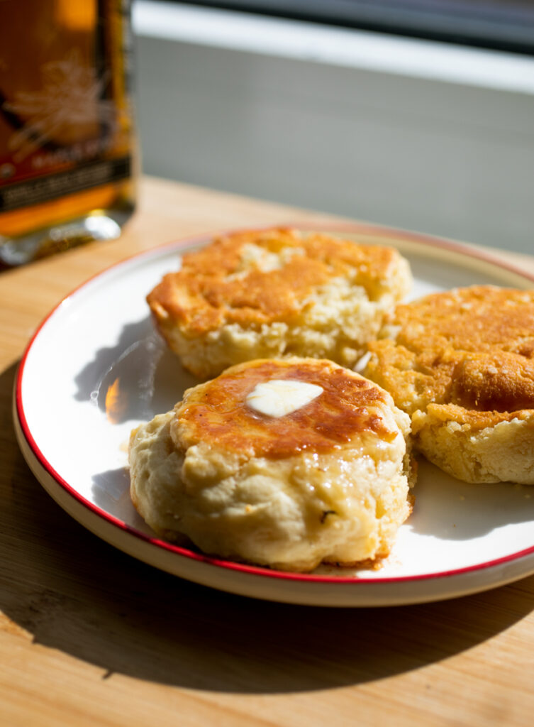 Three thick pancakes with golden surfaces on a white plate with maple syrup in the background.