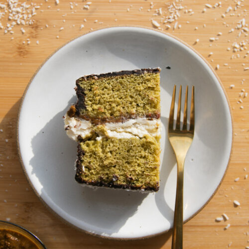 A slice of green pistachio cake with a layer of mascarpone in the center on a white plate with a gold fork.