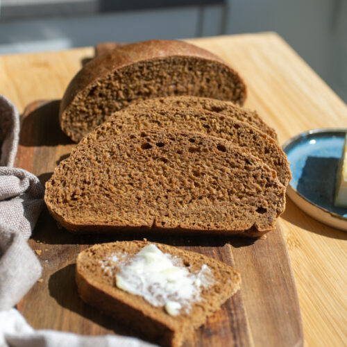 A sliced loaf of whole wheat bread with a buttered slice of bread on a wooden board.
